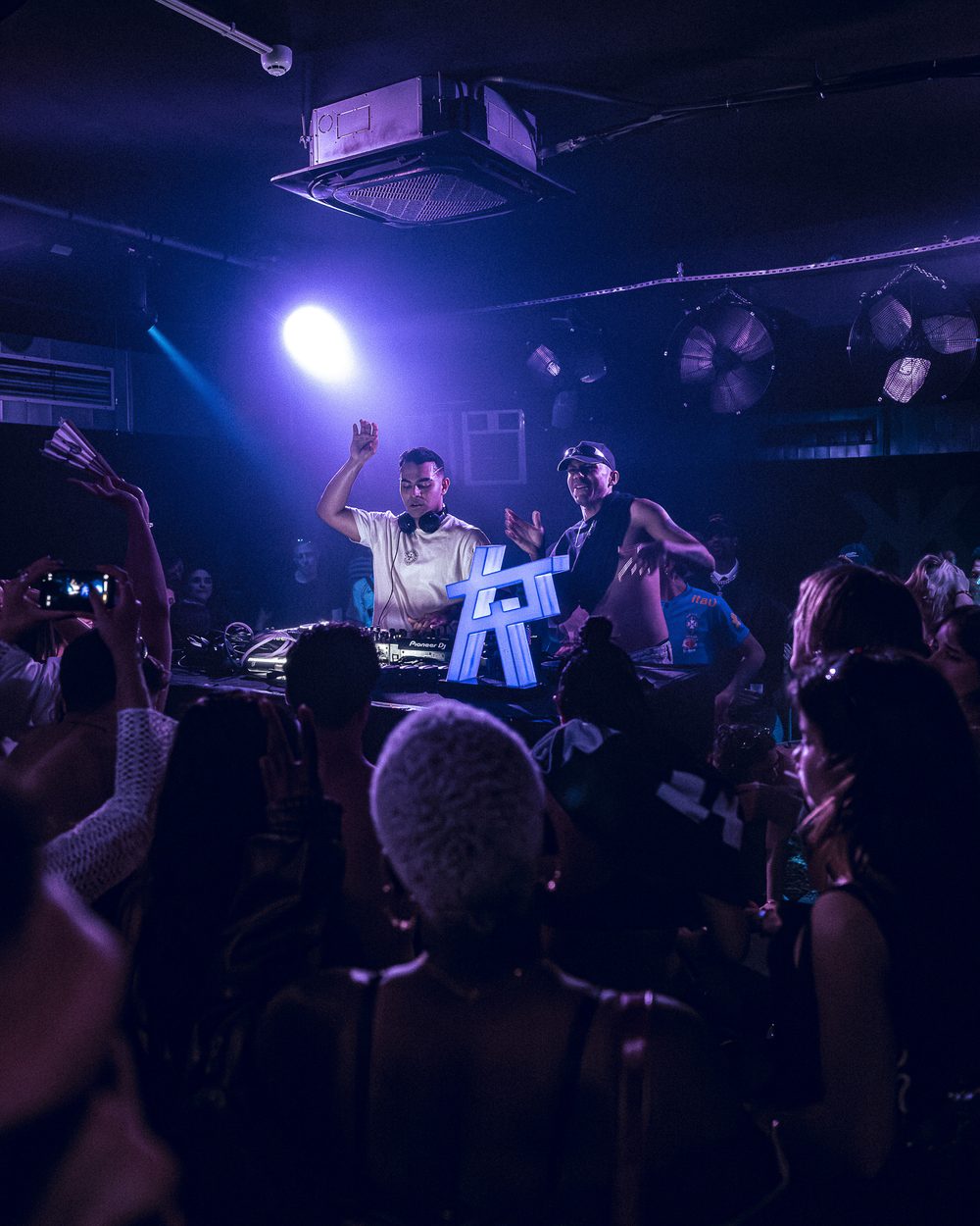 DJ Zauo na cabine, braço no ar, público em luz azul